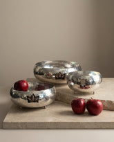 Set of silver metallic bowls with red apples on a stone surface against a beige wall.