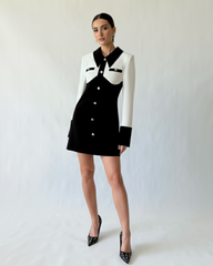 Model wearing black and white crepe mini dress with pearl buttons and tailored silhouette, standing against a white studio backdrop.