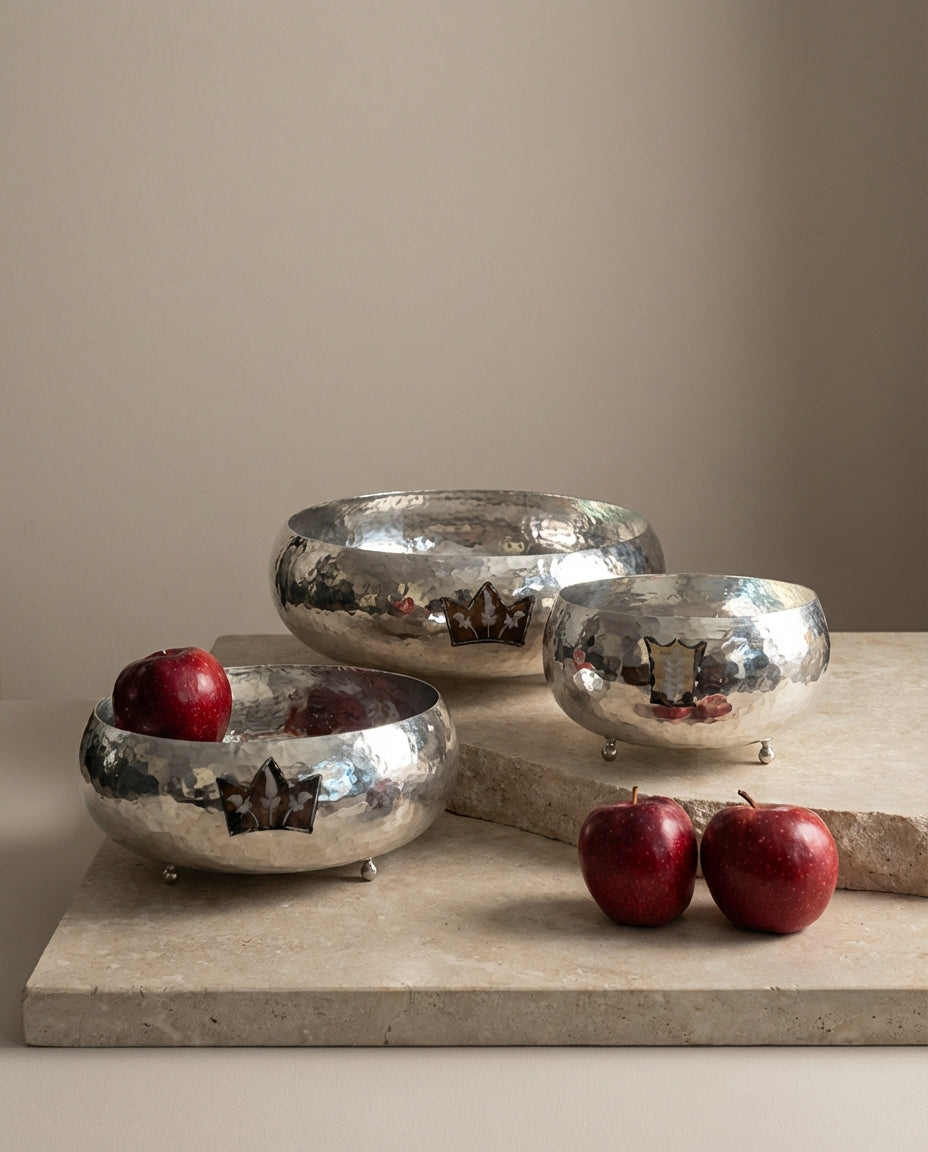 Set of silver metallic bowls with red apples on a stone surface against a beige wall.
