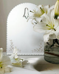 White lilies in a clear vase next to a white card with floral design on a wooden surface.